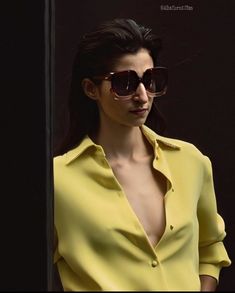 a woman in yellow shirt and sunglasses standing next to a black wall with her hands on her hips