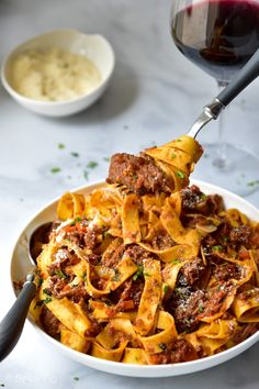 a white bowl filled with pasta and meat next to a glass of wine on a table