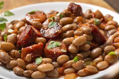 a white plate topped with beans and meats on top of a wooden table next to a green leafy garnish