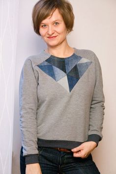 a woman standing in front of a white wall wearing a gray sweater and black jeans