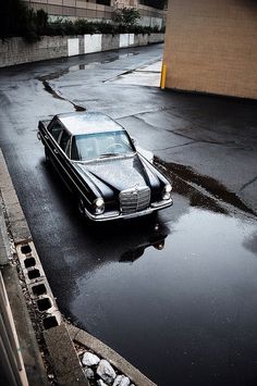 an old black car is parked on the wet street in front of a building and water puddles around it