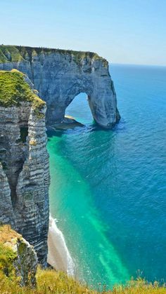 an arch shaped rock formation in the ocean near a beach with green grass and blue water