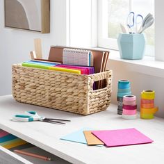 a basket filled with office supplies on top of a white table next to a window