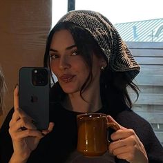 a woman holding up a coffee mug while looking at her cell phone and taking a selfie