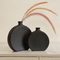 two black vases sitting next to each other on a table with a plant in the background