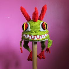 a crocheted green hat with red horns and big eyes on a wooden stick