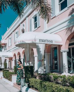 a woman standing in front of a pink building with white awnings and palm trees