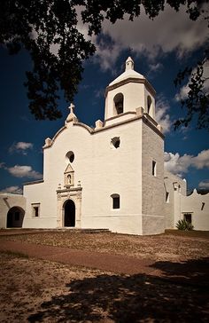 an old white church sits in the middle of a dirt field under a cloudy blue sky