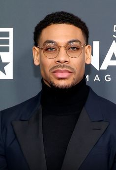 a man wearing glasses and a black turtle neck sweater stands in front of a red carpet