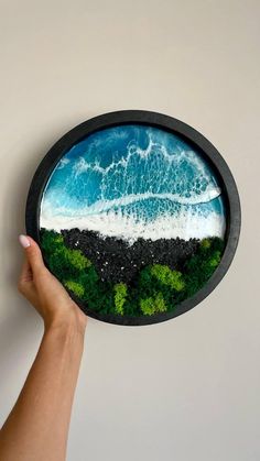 a hand is holding up a painting that looks like water and grass on the beach