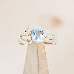 a ring with an aqua blue topaz sits on a wooden stand in front of a beige background