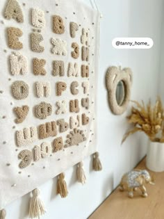 a wall hanging with letters and tassels on it next to a potted plant