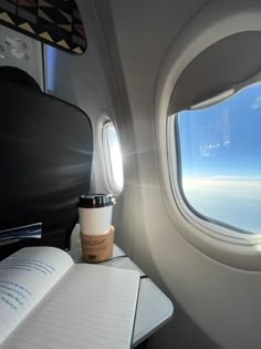 an airplane window with a book and cup on the table next to it, looking out at the sky