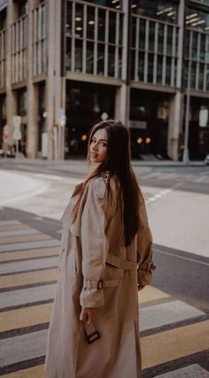 a woman standing on the street in front of a tall building wearing a trench coat