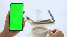 a person holding a cell phone with a green screen in their hand next to a cup of coffee