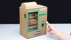 a cardboard vending machine is being held up by a person's hand with a pen in front of it