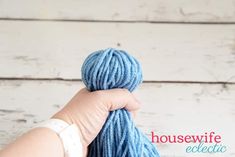 a hand holding a ball of blue yarn in front of a white wooden background with the words housewife eclectic written on it