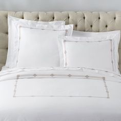 a bed with white sheets and pillows on top of it, in front of a beige headboard