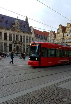 a red and black bus traveling down a street next to tall buildings with people walking on the sidewalk