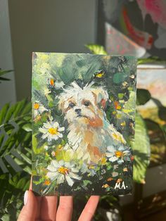 a hand holding up a small painting of a dog with daisies in the foreground