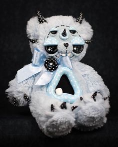 a white teddy bear with blue and black decorations on it's face, sitting against a black background