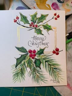 a christmas card with holly leaves and berries on it, sitting next to a plate