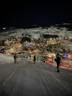 two people walking up the side of a snow covered slope at night with buildings in the background