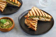 two black plates topped with sandwiches next to a bowl of pesto and pine nuts