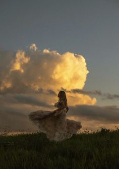 a woman in a long dress standing on top of a lush green field under a cloudy sky