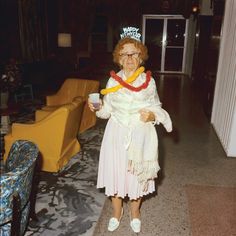 an older woman in a white dress is holding a drink and wearing a tiara