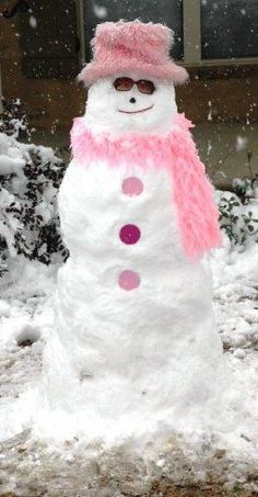 a snowman wearing a pink hat and scarf in front of a house covered in snow