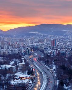 an aerial view of a city at sunset with mountains in the background and cars driving on the road