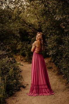 a woman in a pink dress standing on a dirt path surrounded by bushes and trees