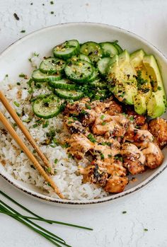 a white bowl filled with rice and chicken next to cucumber, avocado and chopsticks