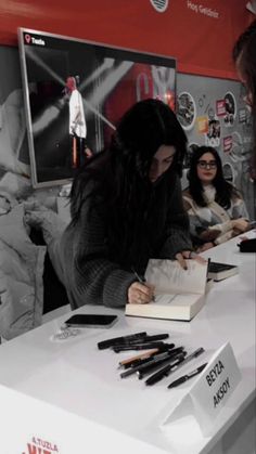 two women are signing books at a book sale with other people in the background and an advertisement on the wall behind them