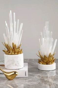 two white marble vases with gold leaf decorations on top of each one, sitting on a table