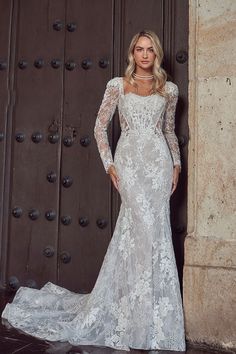 a woman standing in front of a brown door wearing a white wedding dress with long sleeves