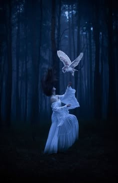 a woman in a white dress holding an owl over her shoulder while standing in the woods