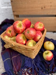 a basket filled with red and green apples on top of a blue cloth next to a wooden crate