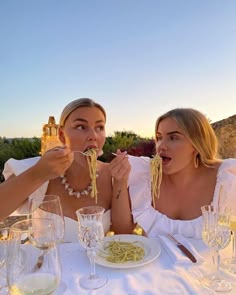 two women sitting at a table eating spaghetti off of their plates with wine glasses in front of them