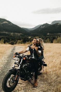 a man and woman sitting on a motorcycle in the middle of a field with mountains behind them