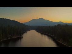 an aerial view of a lake surrounded by trees and mountains in the distance at sunset