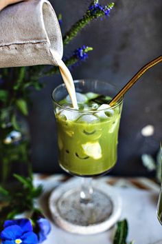 a glass filled with green liquid being poured from a pitcher into a cup next to blue flowers