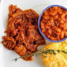 a white plate topped with meat and beans next to biscuit on top of a table