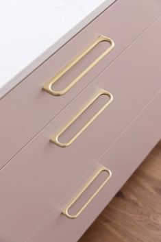 a pink dresser with brass handles on it's drawers and wood flooring in the background