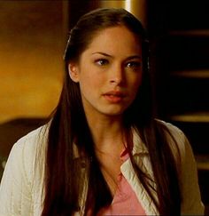 a woman with long brown hair wearing a white cardigan and pink shirt looking at the camera