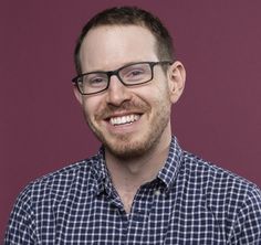 a man with glasses smiling at the camera in front of a purple wall and wearing a blue checkered shirt