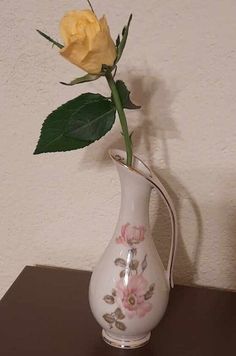 a single yellow rose in a white vase on a brown table next to a wall