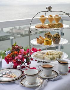 a table topped with plates and cups filled with desserts next to the ocean on top of a balcony