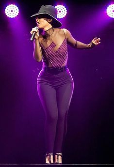 a woman standing on top of a stage wearing purple pants and a hat with her hands out